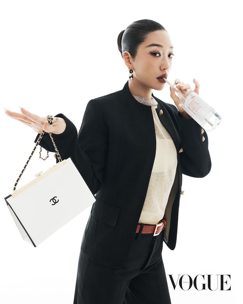 Woman in dark suit jacket poses with white Chanel purse and water bottle against a white backdrop, styled hair and dark red lipstick.