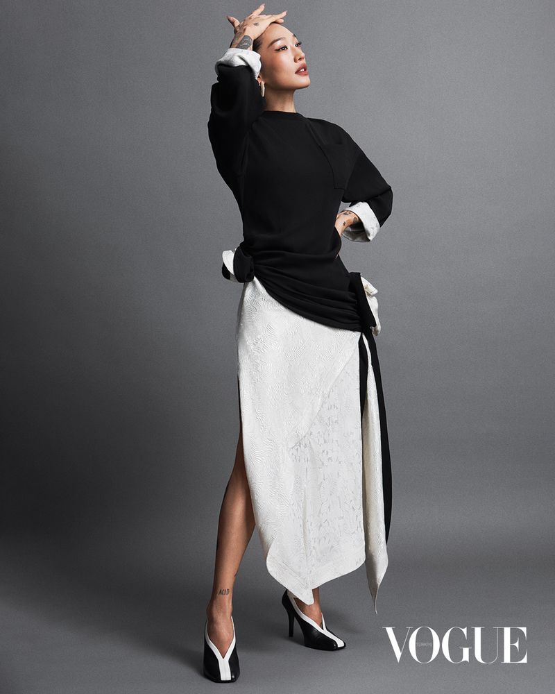 Elegant adult woman poses in black top and white skirt with matching shoes, set against a neutral gray backdrop in a studio shot.