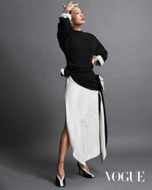 Elegant adult woman poses in black top and white skirt with matching shoes, set against a neutral gray backdrop in a studio shot.