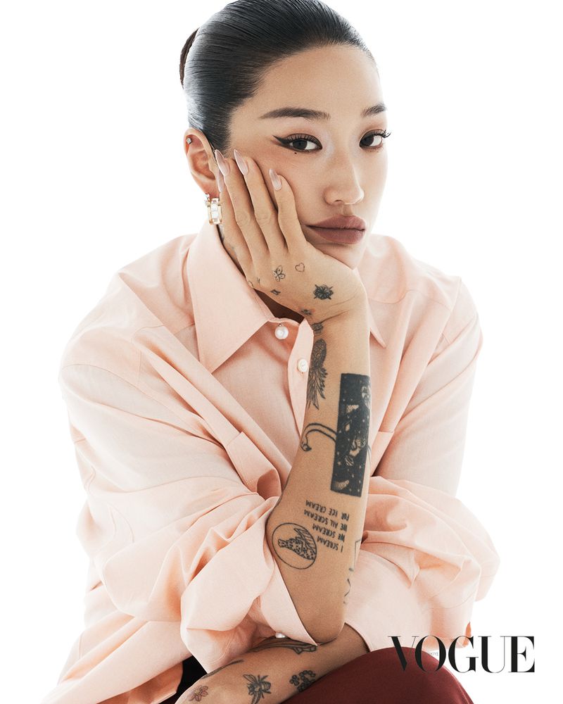 Portrait of an adult Asian woman with tattoos and a pink shirt sitting in a studio in front of a white background.