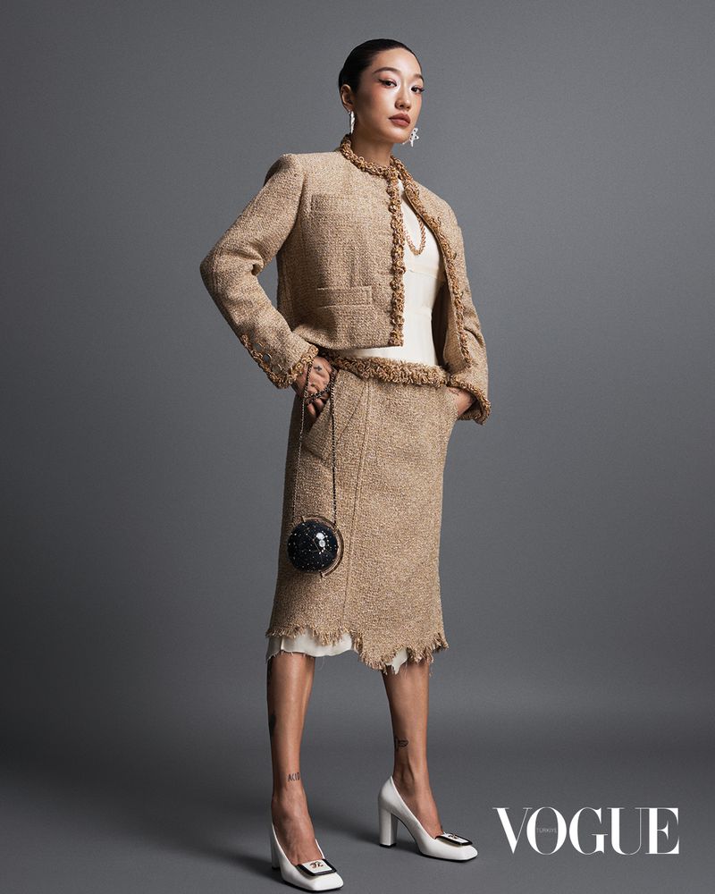 An Asian woman poses in a tan tweed skirt suit, a white top, and white high heels, carrying a disco ball purse in a studio shot.