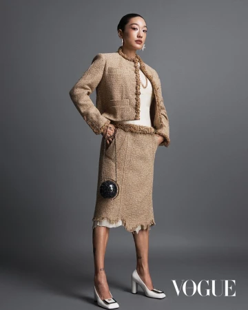 An Asian woman poses in a tan tweed skirt suit, a white top, and white high heels, carrying a disco ball purse in a studio shot.