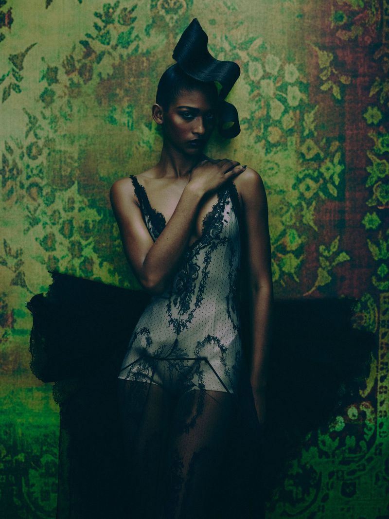 Woman poses in black lace lingerie and elaborate headwear in front of floral wallpaper in a studio shot.