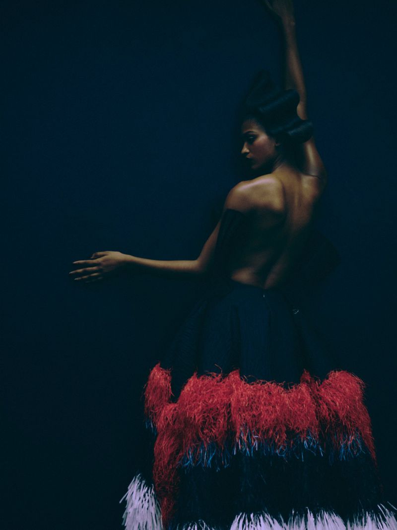 Woman in a feather skirt, posed with arms raised against a dark backdrop. The model's back is bare, showing a serene and artistic studio shot.