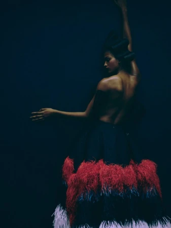 Woman in a feather skirt, posed with arms raised against a dark backdrop. The model's back is bare, showing a serene and artistic studio shot.