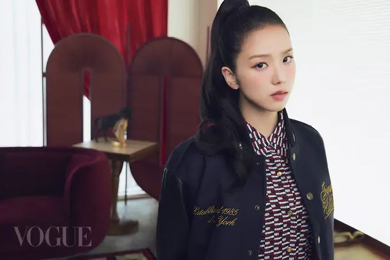 A portrait of a woman with dark hair in a high ponytail, wearing a letterman jacket in an indoor setting, with a red curtain backdrop.