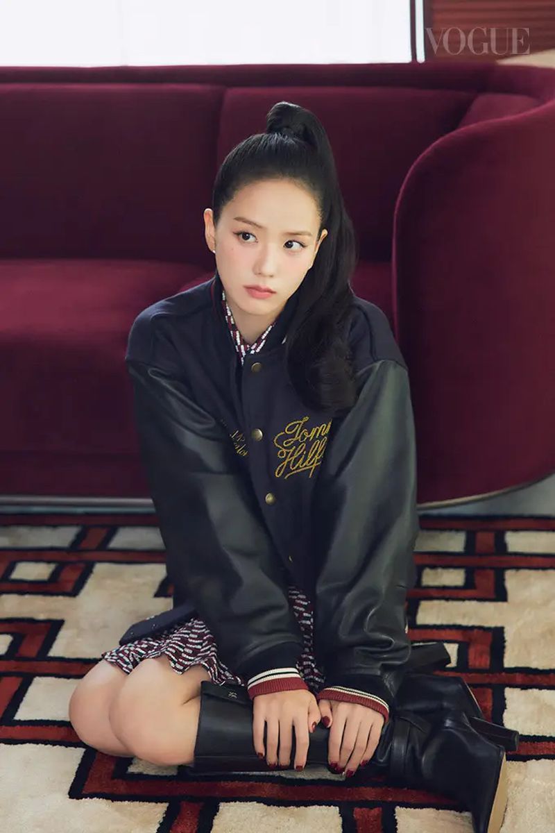 Portrait of a young Asian woman with dark hair in a high ponytail wearing a Tommy Hilfiger jacket and boots while sitting on a patterned carpet.