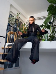 Model wearing a black sheer blouse and wide trousers sitting high on an indoor ledge surrounded by potted plants.