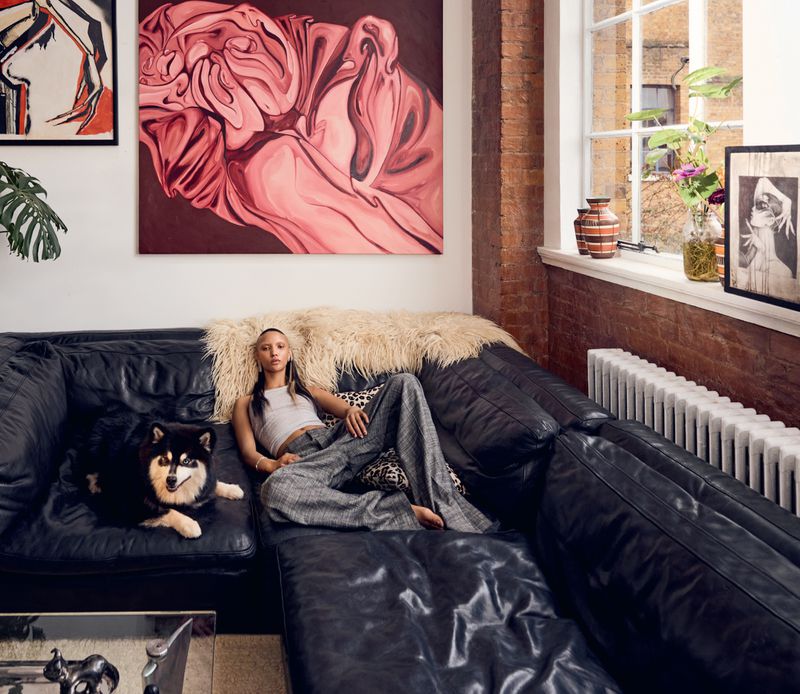 Woman lounges on a black leather couch with her dog, paintings and exposed brick add character to the indoor living space.