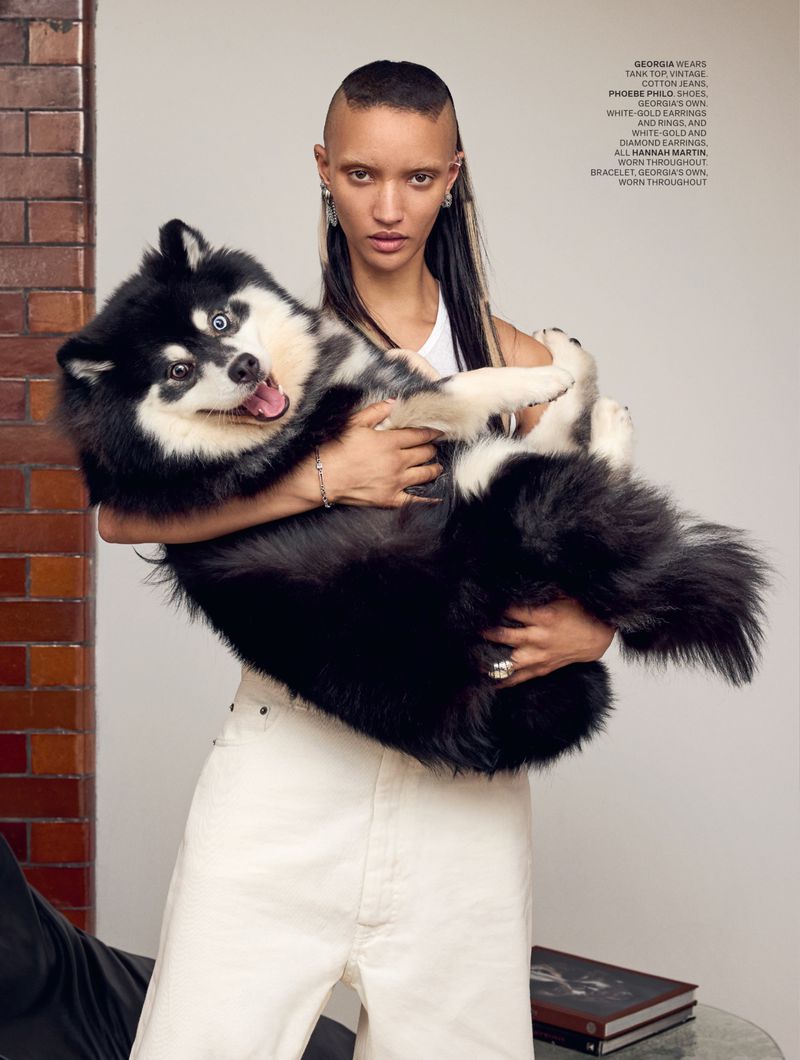 A woman holding a large black and white husky in a fashion editorial shot. The model is wearing a white top and white jeans.