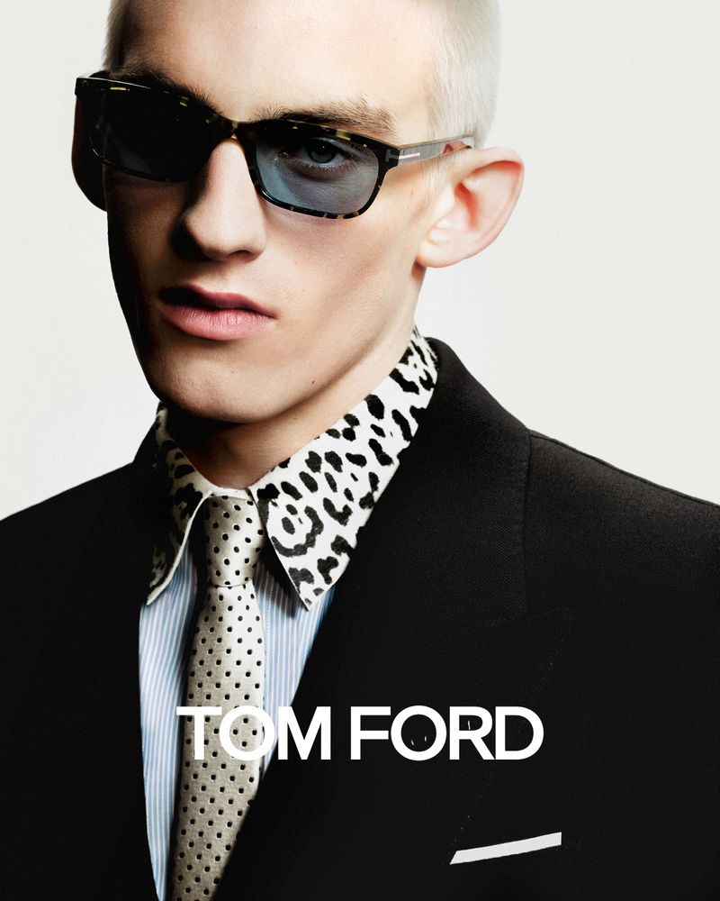 A fair-skinned adult male models sunglasses, wearing a black suit, spotted tie, and collared shirt against an off-white background.