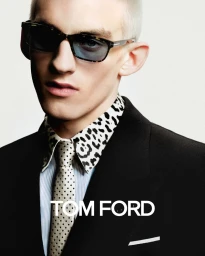 Close-up portrait of a male model wearing tortoiseshell sunglasses, a black suit, leopard-print collar shirt, and polka-dot tie.