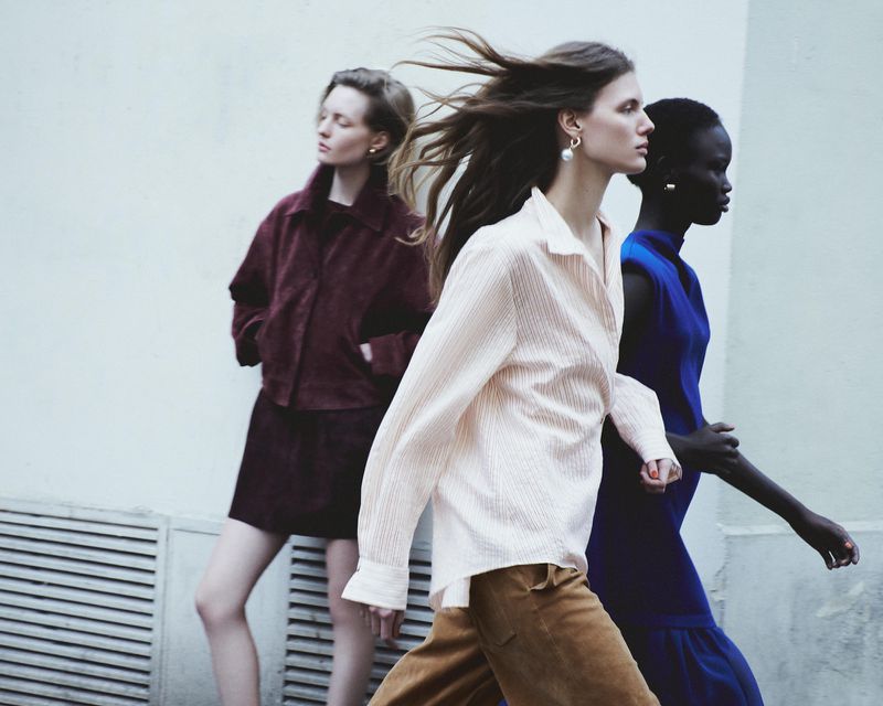 Three fashion models walk in front of a white wall. Two are in profile, one looks left, in an outdoor fashion shot with diffused lighting.