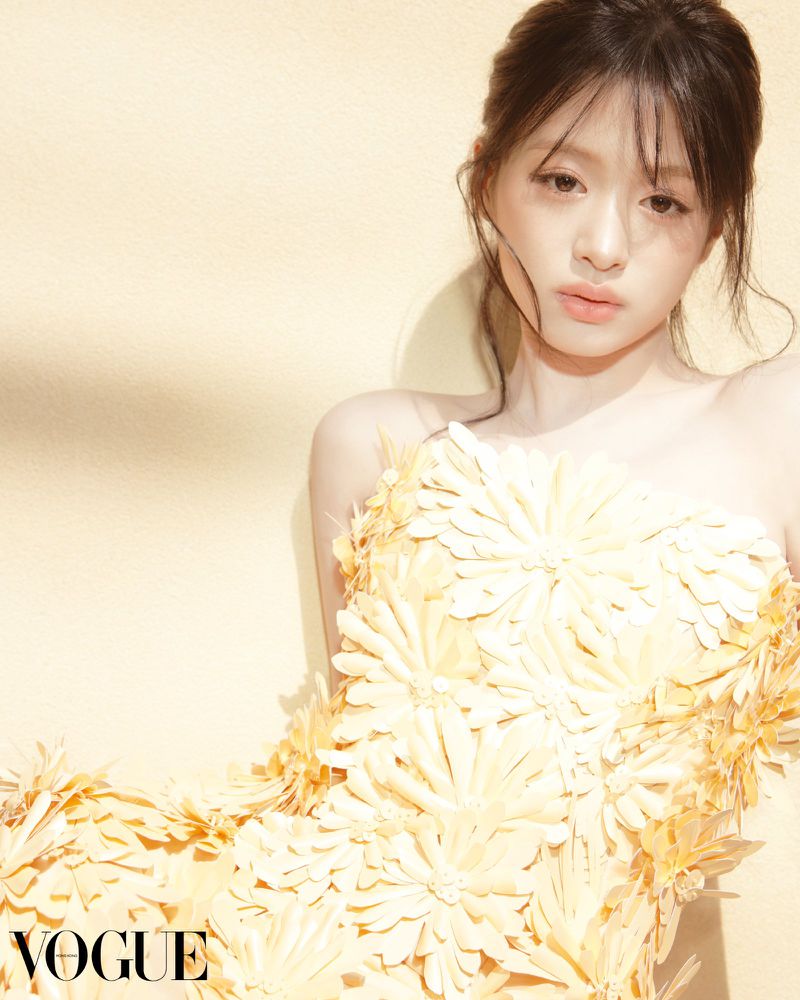 Close-up portrait of an adult female model in strapless yellow flower dress, against a light yellow background for Vogue magazine.