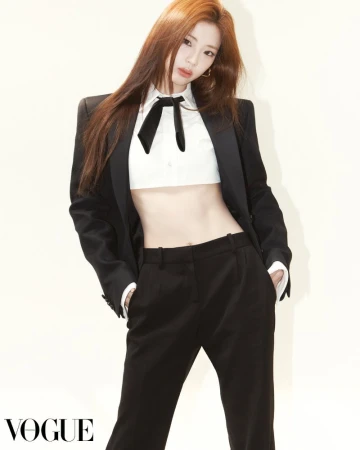 Fashion portrait of a woman with long auburn hair wearing a black blazer, black pants, and a white cropped shirt with a black bow tie.