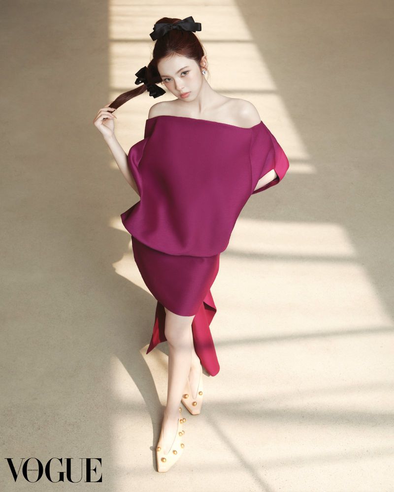 A woman poses in a magenta off-shoulder dress and cream heels on a beige surface, accessorized with black ribbon hair ties, shot at eye-level.