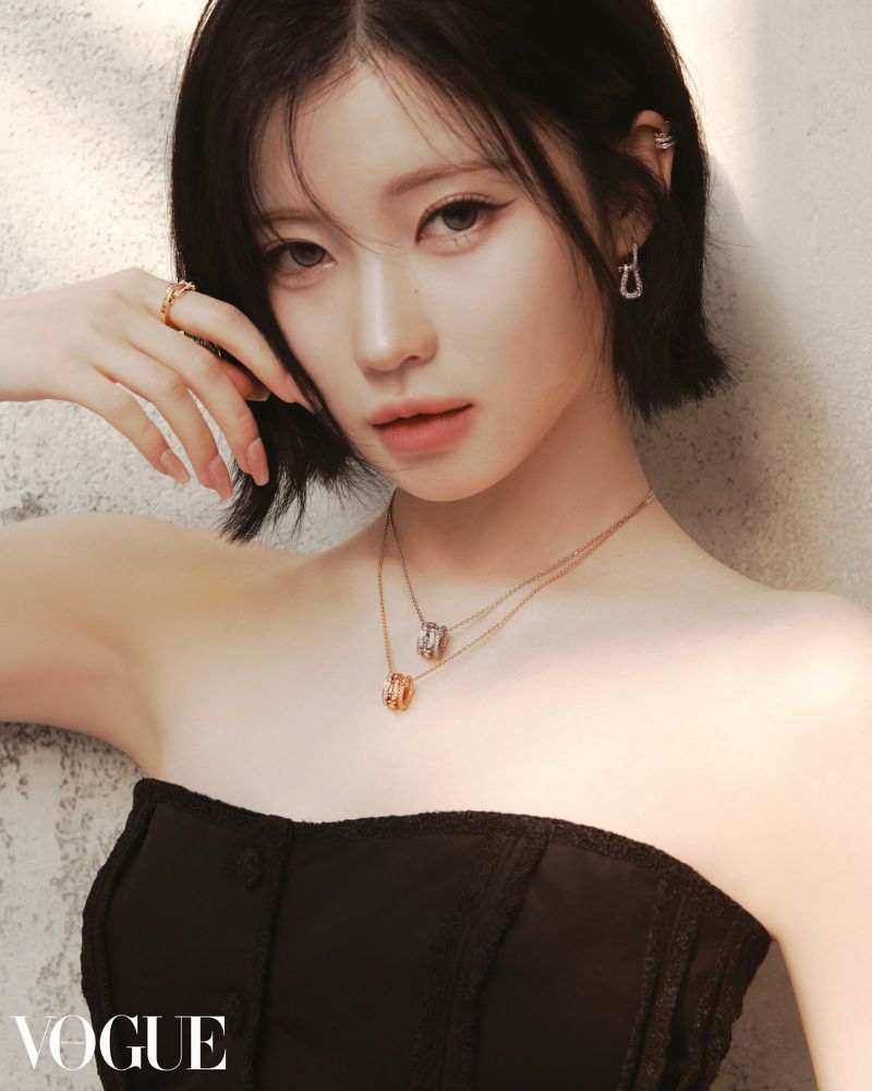 Portrait of a young Asian woman with short dark hair wearing a black strapless top and gold jewelry, posing in front of a textured wall.