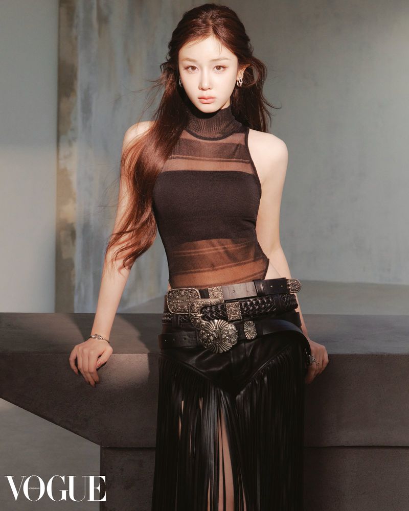 Elegant woman in black semi-sheer top, multiple belts, and fringed skirt poses indoors with soft lighting and Vogue logo.