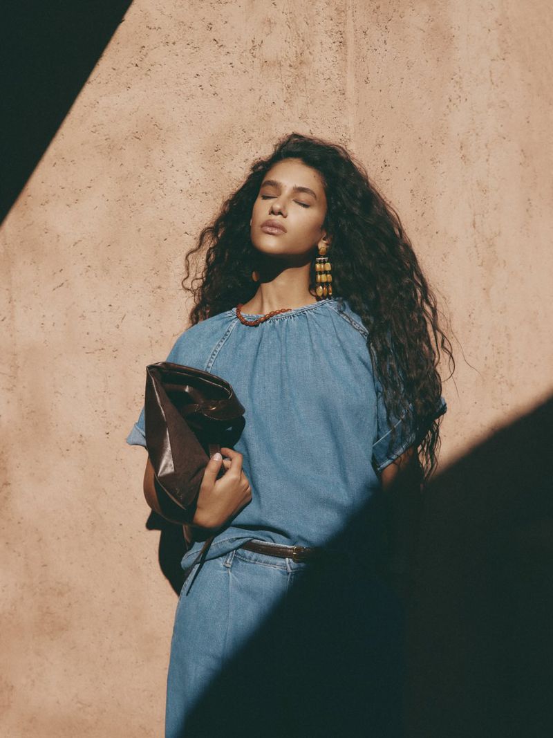 Portrait of a woman with long curly hair in a denim dress, holding a leather clutch, bathed in sunlight against a textured wall.
