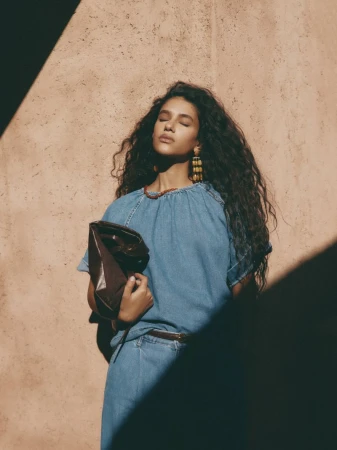 Portrait of a woman with long curly hair in a denim dress, holding a leather clutch, bathed in sunlight against a textured wall.