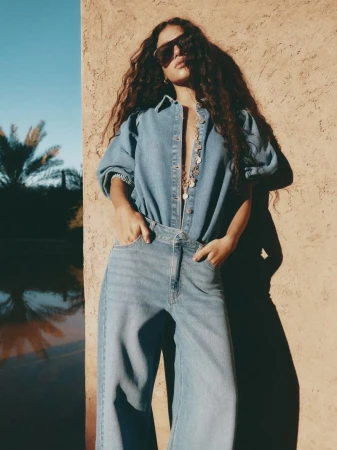 A woman with long curly hair poses in a denim jumpsuit and sunglasses, standing in front of a textured wall on a sunny day.