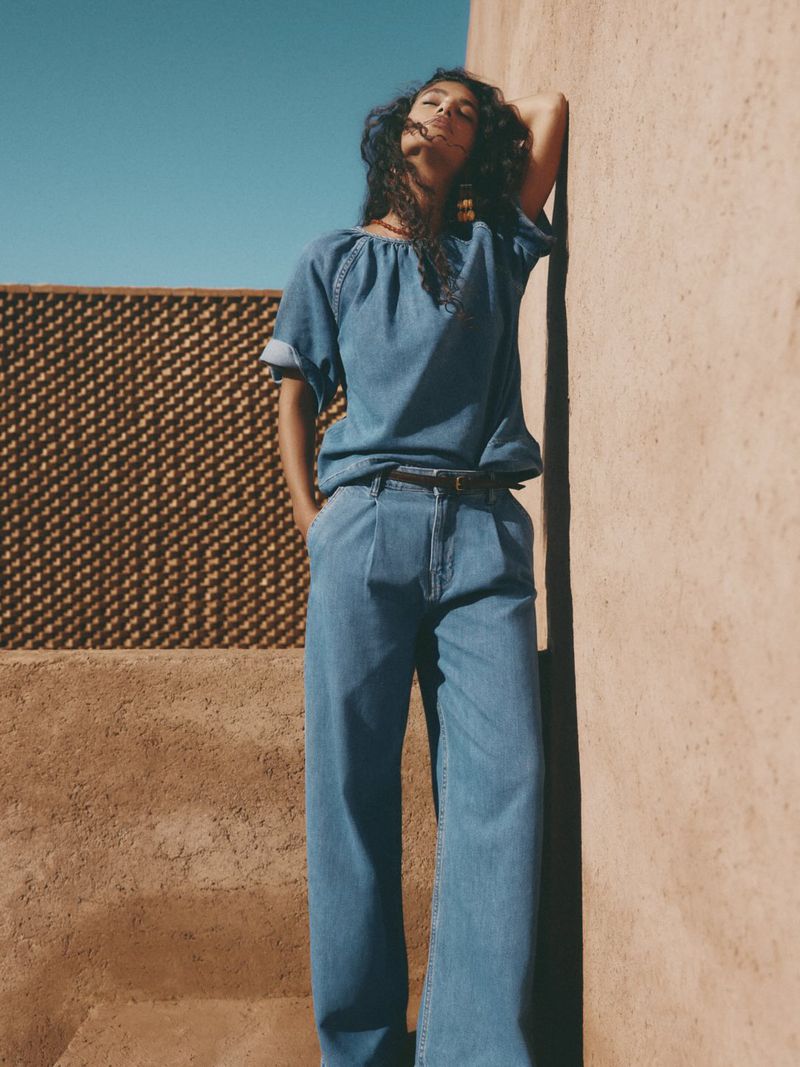 Woman in denim blouse and wide-leg jeans poses against earthen wall under a clear sky in an outdoor setting, fashion photography.