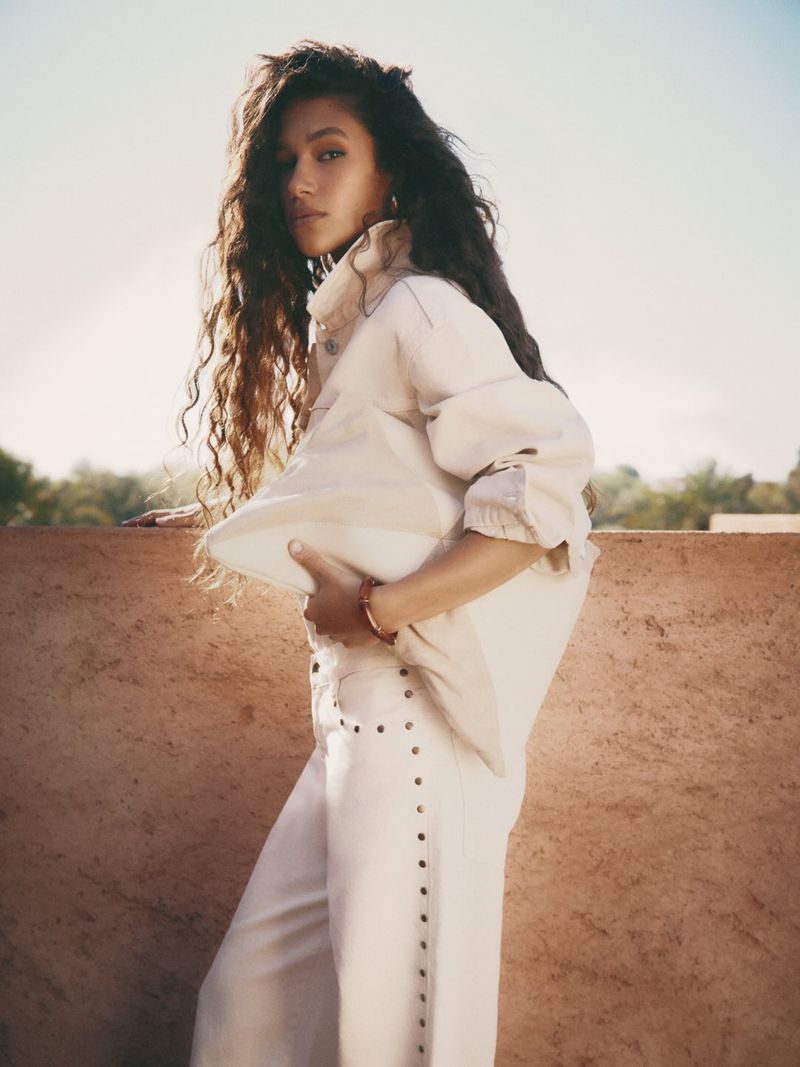 Stylish woman with long curly hair poses in a beige jacket and pants against an outdoor background with a sky view.  
