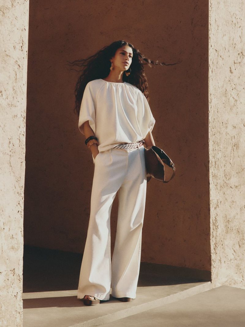 A fashionable woman with long curly hair stands in a white blouse and pants, complemented by a brown purse, captured in medium shot.