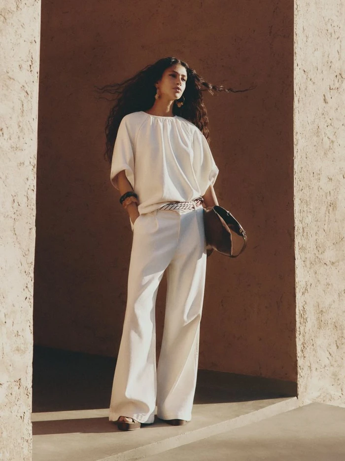Model with long dark hair wearing a white top and trousers holding a handbag within earthy textured stone architecture.