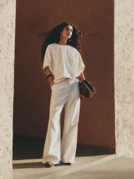 Model with long dark hair wearing a white top and trousers holding a handbag within earthy textured stone architecture.