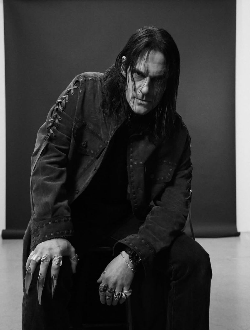 Monochrome portrait of a man with long black hair, wearing a dark jacket and multiple rings, including claw-like finger adornments, seated indoors.