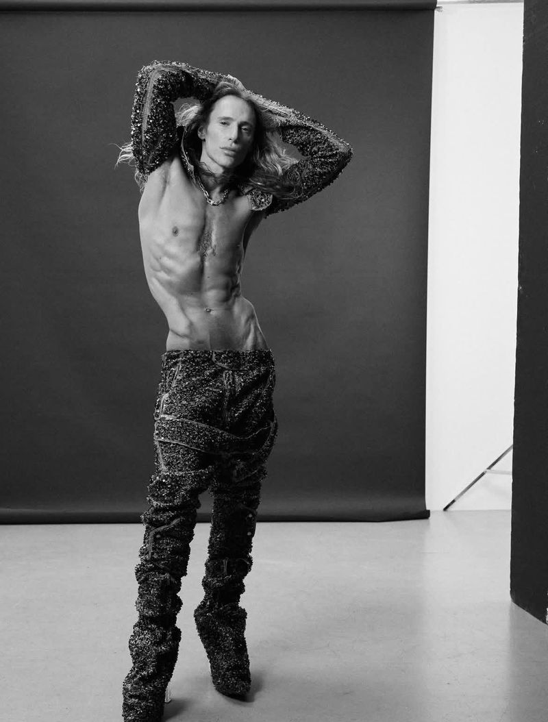 Striking black and white portrait of a slender, shirtless adult male in embellished pants and top, posing against a dark backdrop in a studio.