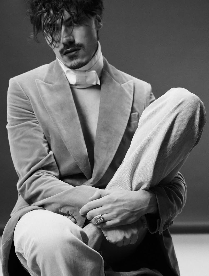 Black and white portrait of a man in a suit sitting down with hands clasped, showcasing fashion and style in a simple background.