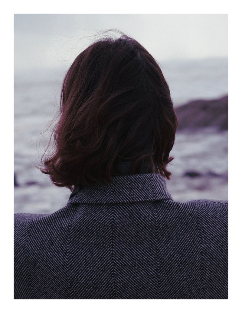 Back view of adult woman with short brunette hair wearing a herringbone jacket, facing the ocean under an overcast sky.