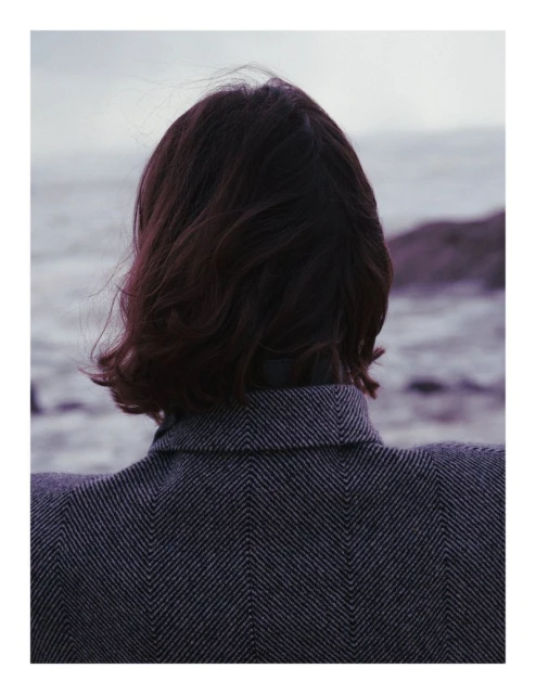 Back view of adult woman with short brunette hair wearing a herringbone jacket, facing the ocean under an overcast sky.