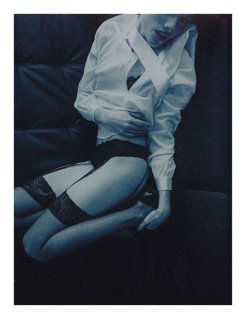 Woman in unbuttoned shirt, black lingerie, and stockings kneeling on dark fabric. Dark, moody lighting creates a sensual, intimate scene.