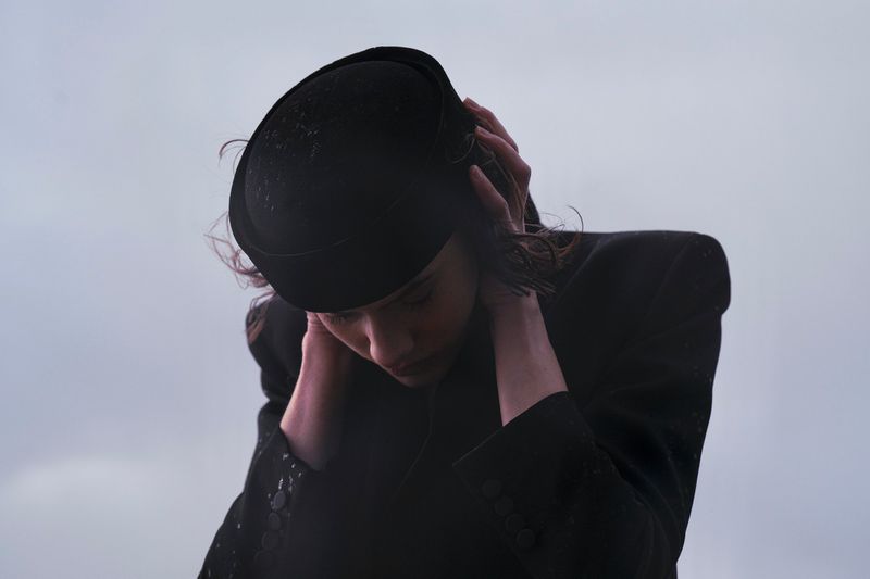 Woman wearing a black hat and coat with her hands on her neck, looking down in front of a light grey background.