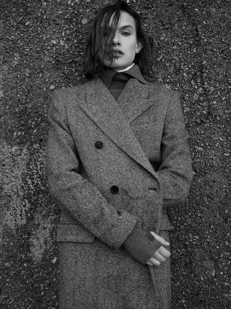Black and white portrait of an adult female in a gray herringbone coat, looking directly at the camera with a textured background.