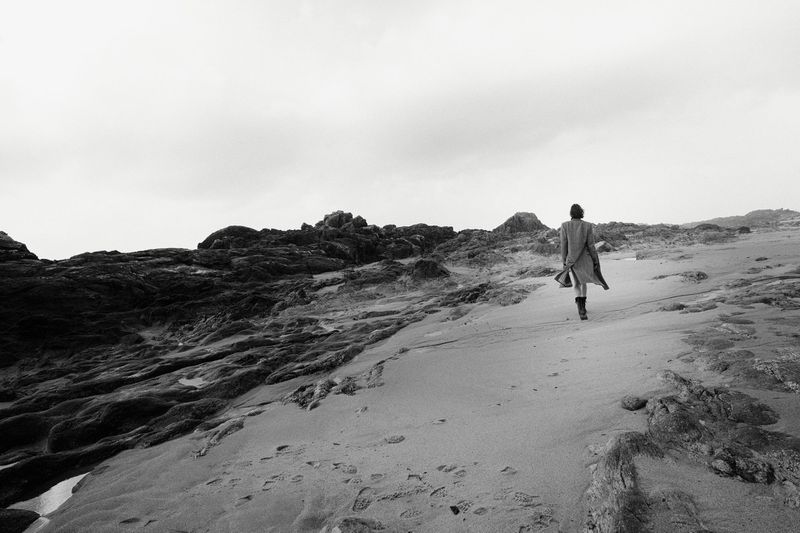 Person in coat walks on sandy beach with dark rocks under cloudy sky, creating a desolate and windswept scene in black and white.