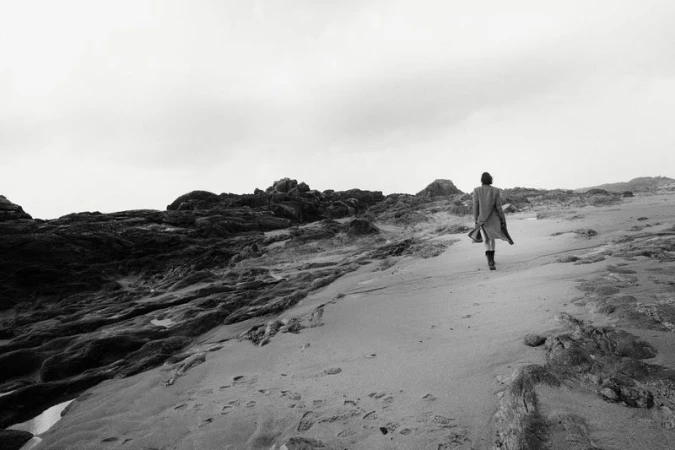 Person in coat walks on sandy beach with dark rocks under cloudy sky, creating a desolate and windswept scene in black and white.