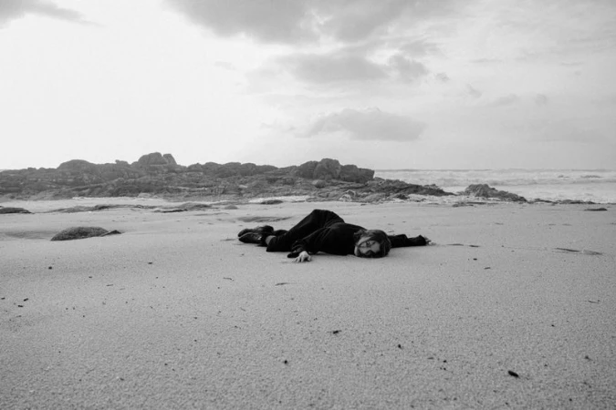 Model lying on a sandy beach wearing dark clothing against a rocky coastline under an overcast sky.