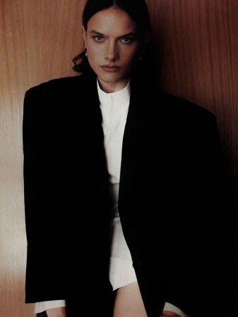 Model wearing an oversized black blazer over a white high-neck shirt against a warm wood panel wall.