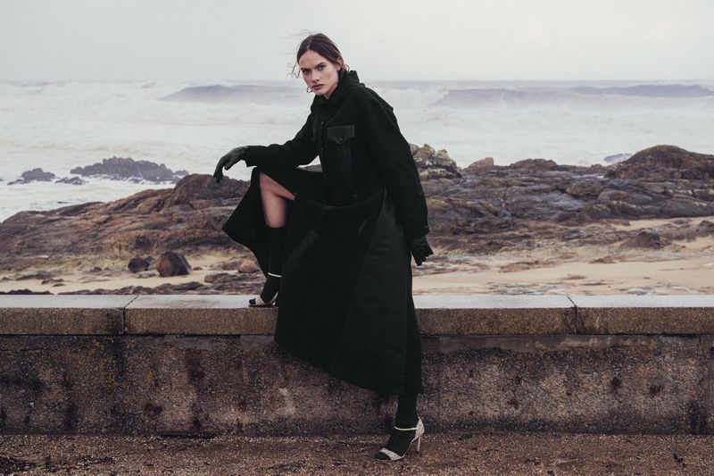 A stylish woman in a dark coat sits on a low wall with a sea and rocky shoreline in the background, posing for a fashion editorial shoot.