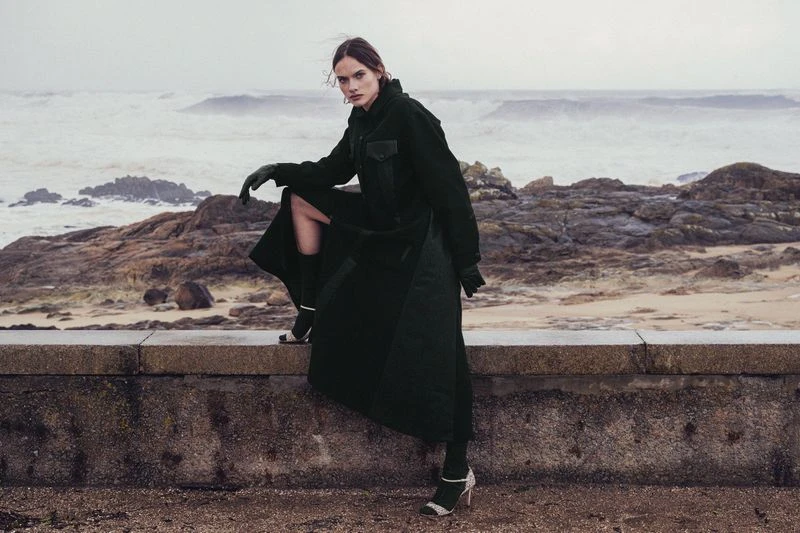 A stylish woman in a dark coat sits on a low wall with a sea and rocky shoreline in the background, posing for a fashion editorial shoot.