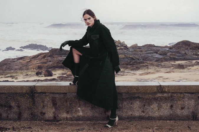 A stylish woman in a dark coat sits on a low wall with a sea and rocky shoreline in the background, posing for a fashion editorial shoot.