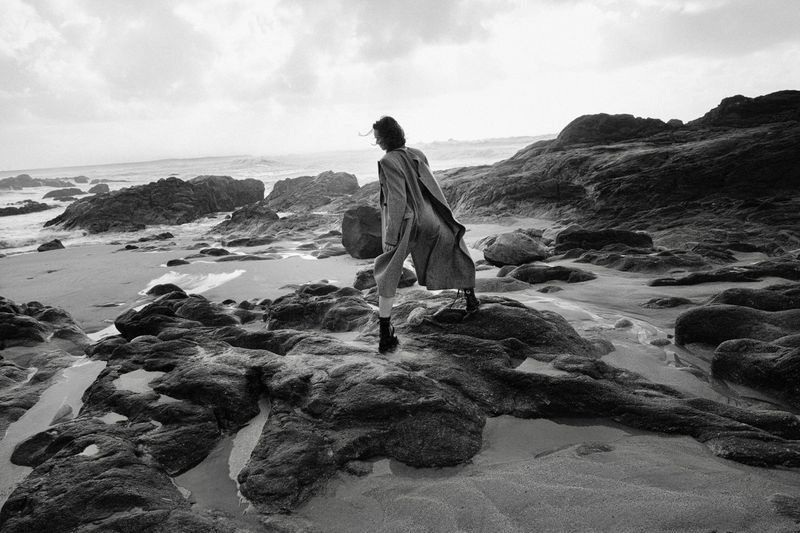 Monochrome fashion photograph of a model in a long coat standing on wet rocks along a rocky coastline with ocean waves.