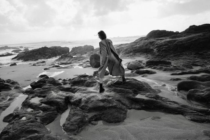 Monochrome fashion photograph of a model in a long coat standing on wet rocks along a rocky coastline with ocean waves.