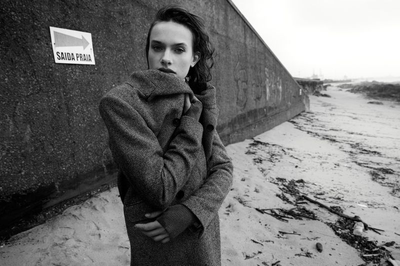 Monochrome outdoor fashion portrait of a young adult female in a coat standing on a sandy beach next to a concrete barrier.
