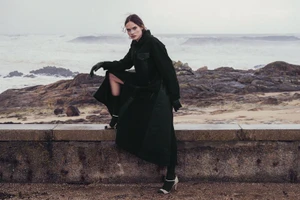 Model in a long black coat crouching on a stone wall by a rocky coast with choppy ocean waves in the background.
