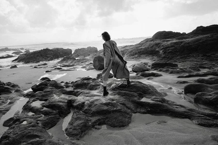 Monochrome fashion photograph of a model in a long coat standing on wet rocks along a rocky coastline with ocean waves.
